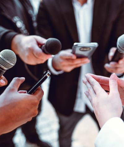 A group of reporters hold up microphones and cellphones, reporters and interviewees faces are obscured.