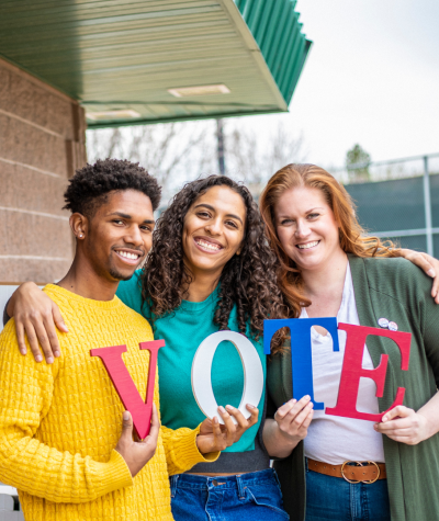 A picture of three people holding letters that spell VOTE. They are standing close together in front of a polling location.