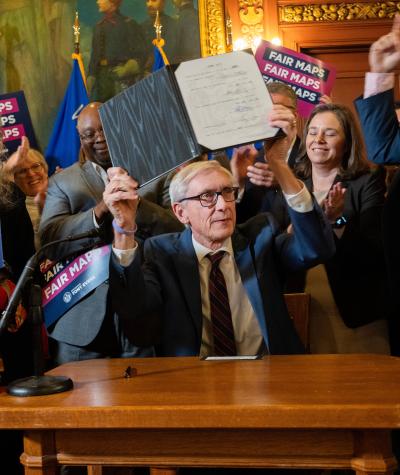 Tony Evers seated at a desk holding documents over his head surrounded by people cheering and holding signs
