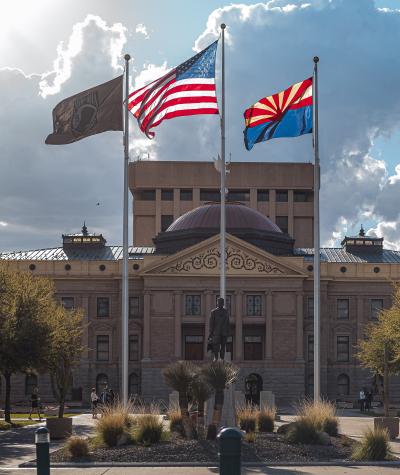 The Arizona state flag with the U.S. flag flying in front of a building with palm trees in the background