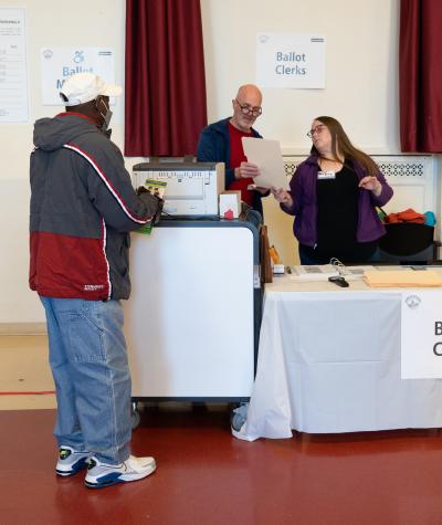 A woman and a man examine a piece of paper while standing next to a machine. A man stands facing them. A sign on the wall says "Ballot Clerks"