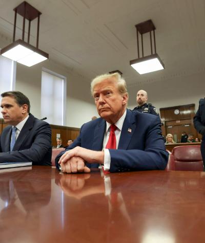 Donald Trump seated at a large table with another man beside him and a guard standing behind him