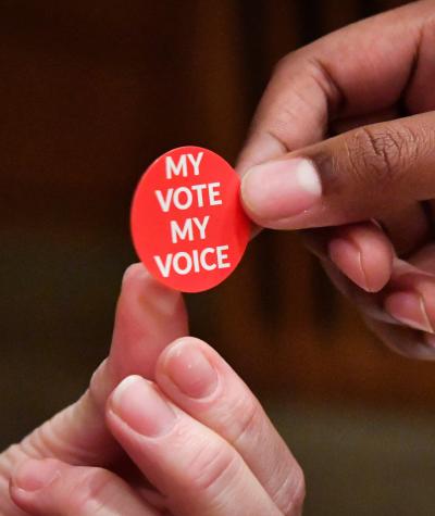 Close up of a person handing a red sticker which says "My Vote, My Voice" to another person.
