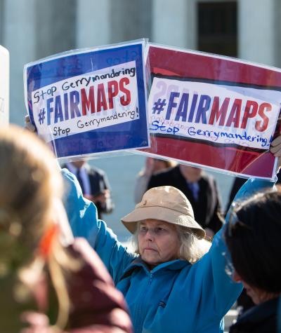 A woman standing in a crowd holds two signs that say "#FAIRMAPS"