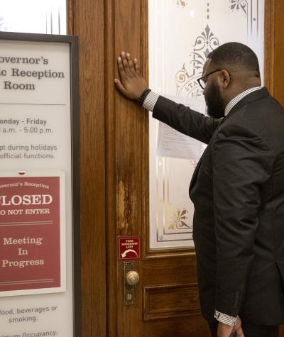A man stands with his hand against a door. A sign on the doors reads "Governor's Public Reception Room, Closed, Do Not Enter, Meeting in Progress"