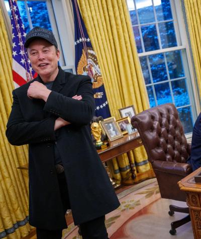 Elon Musk standing up with a smile on his face while Donald Trump sits behind him at his desk in the Oval Office