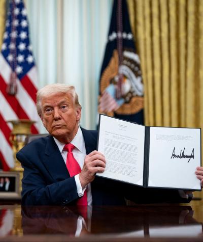 Donald Trump sitting at a desk in the Oval Office holding up an executive order with his signature on it.