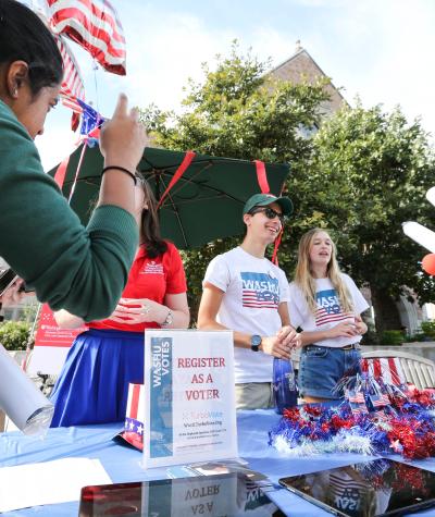 Two smiling people stand behind a table with a sign that says "Register as a voter". Two other people are coming up to the table.
