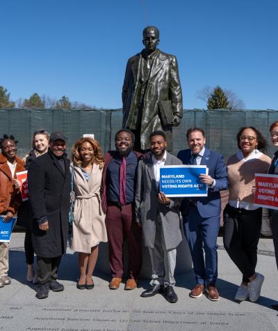 A group of people standing in front of a statue holding signs that say "Maryland needs a voting rights act"