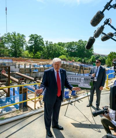 Trump standing in front of a construction site talking to a group of reporters with microphones and cameras.