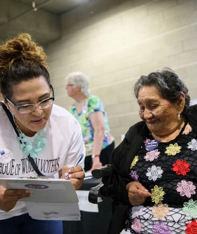 A woman lean fills out a paper on a clipboard next to another woman looking on who is holding a small American flag