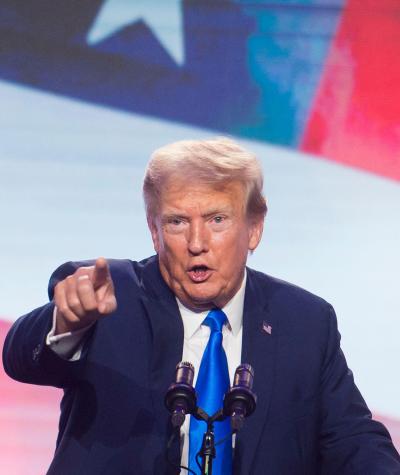Donald Trump speaking while pointing at the camera in front of a giant American flag
