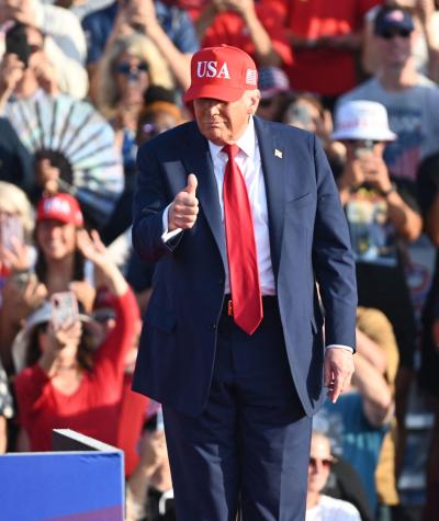 Donald Trump wearing red tie and usa hat giving thumbs up to a crowd of people.