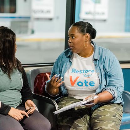 A person wearing a Restore Your Vote t-shirt talking to another person on a bench.