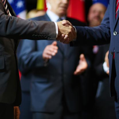 Two men in suits shaking hand with people clapping in the background