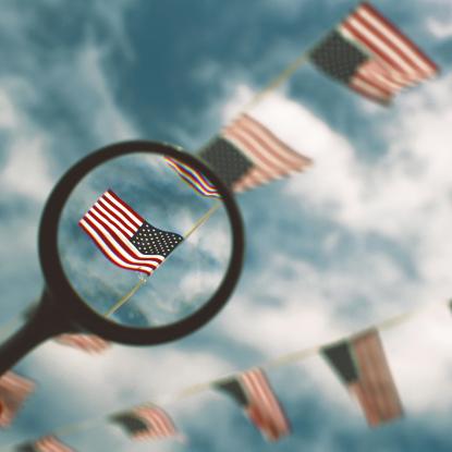 A magnifying glass highlighting one American flag out of a string of them hung up in front of a blue sky with white clouds