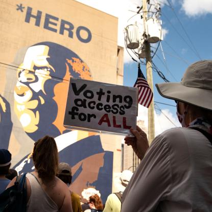 A woman holds a sign which says "Voting Access for All" while standing in front of a large mural of John Lewis.