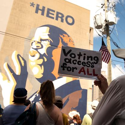 A woman holds a sign which says "Voting Access for All" while standing in front of a large mural of John Lewis.