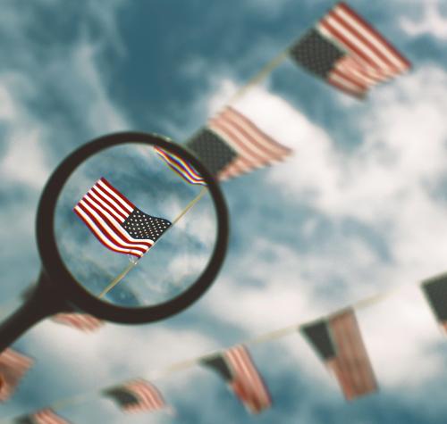 A magnifying glass highlighting one American flag out of a string of them hung up in front of a blue sky with white clouds