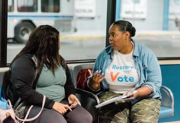 A person wearing a Restore Your Vote t-shirt talking to another person on a bench.