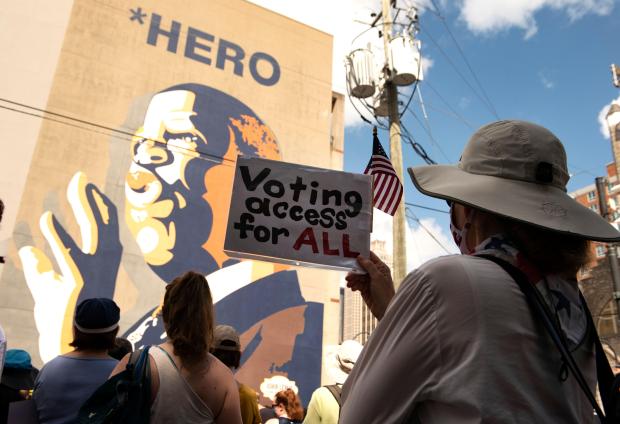 A woman with her back to the camera holds a sign which says "Voting access for ALL", while standing with others in front of a large mural of John Lewis.