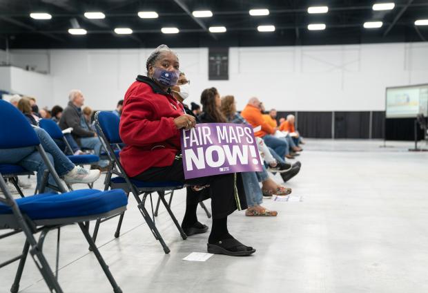 A woman seated in the front row of a group of folding chairs holds a sign which reads "Fair maps now".