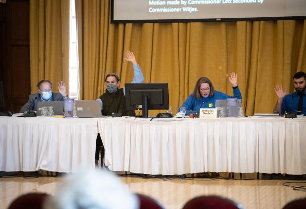 People seated behind a table draped with a white tablecloth raise their hands.