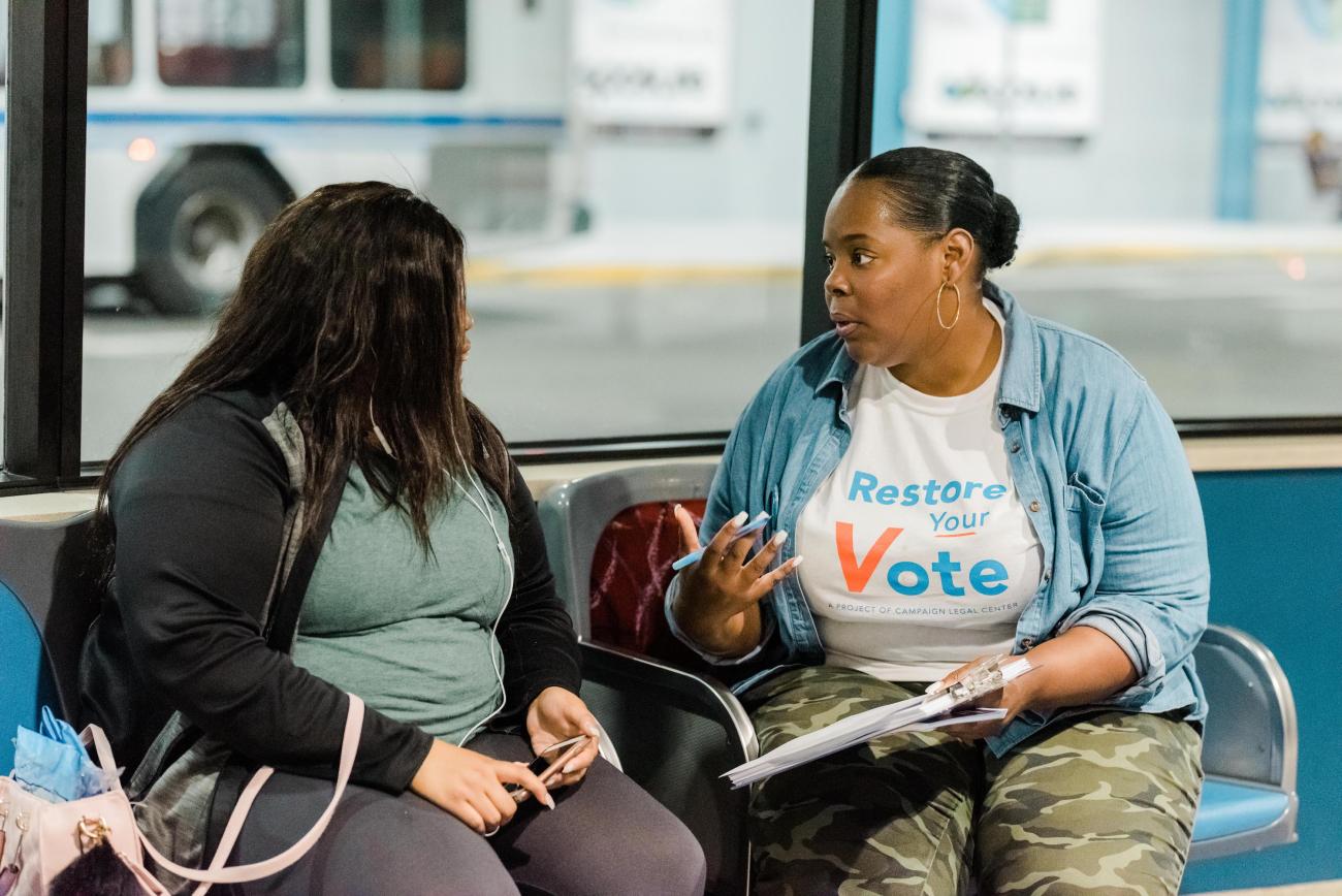 A person wearing a Restore Your Vote t-shirt talking to another person on a bench